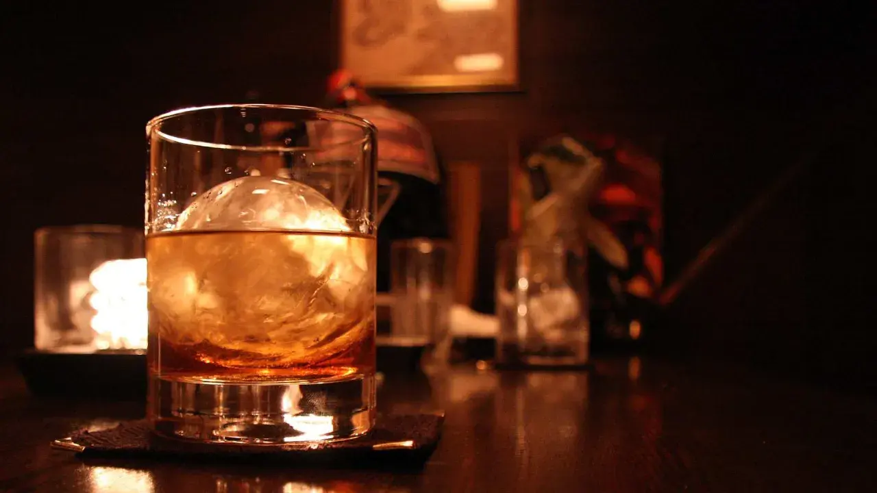Ein Glas bernsteinfarbener Whisky mit einem großen Eisball. Im Hintergrund sind weitere Gläser und eine gedämpfte Beleuchtung zu sehen, die zum Genuss einlädt, wenn man Whisky mit Eis trinkt.
