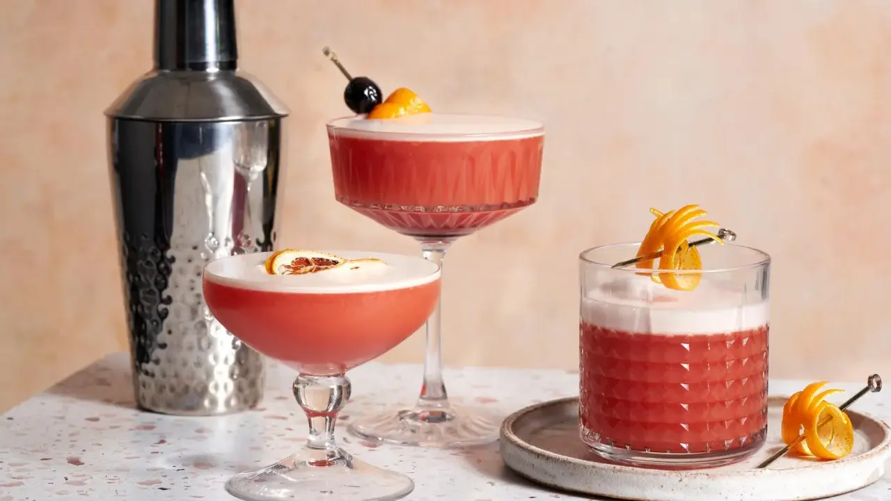 Drei erfrischende Campari Sours, garniert mit Orange und Kirsche, neben einem Cocktail-Shaker.