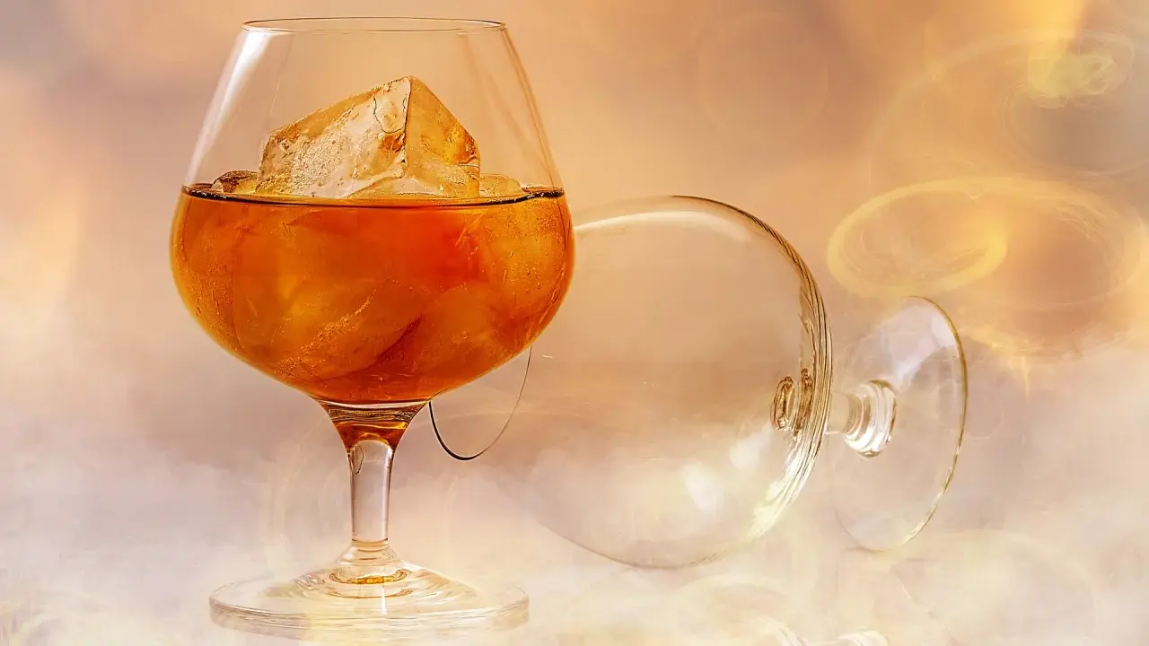 Ein Glas bernsteinfarbener Brandy mit Eiswürfeln, daneben ein leeres, umgekipptes Glas. Man fragt sich, wie schmeckt brandy.