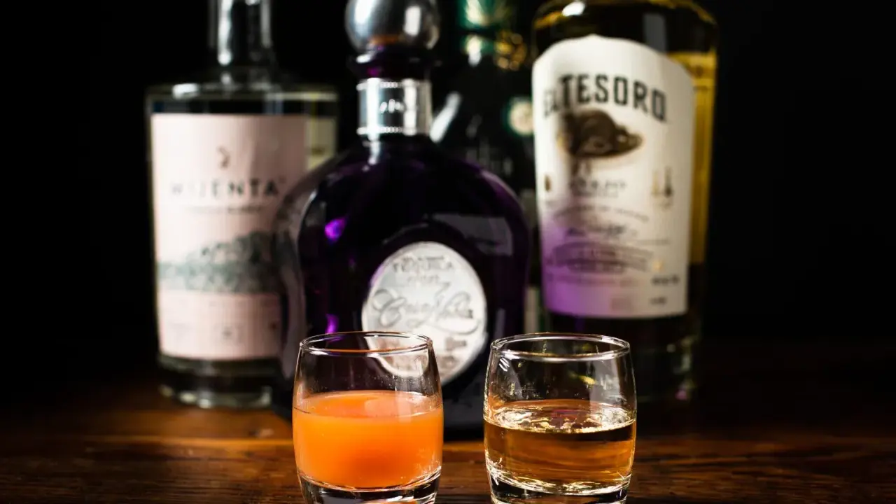 Zwei Gläser mit Tequila und einem orangen Getränk, bereit, um sangrita selber machen zu üben. Flaschen im Hintergrund.