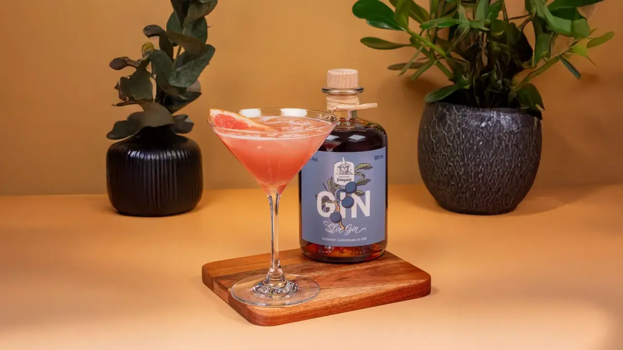 Ein Glas mit pinkfarbenem Gin-Cocktail, garniert mit Grapefruit, neben einer Flasche Sloe Gin. Dekorative Pflanzen im Hintergrund.