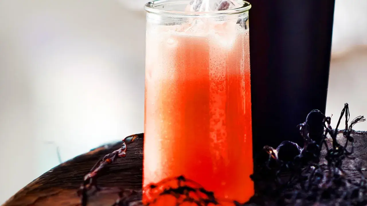 Erfrischender Aperol Sour mit Eiswürfeln in einem Glas, daneben ein dunkler Becher und Spinnweben-Deko.