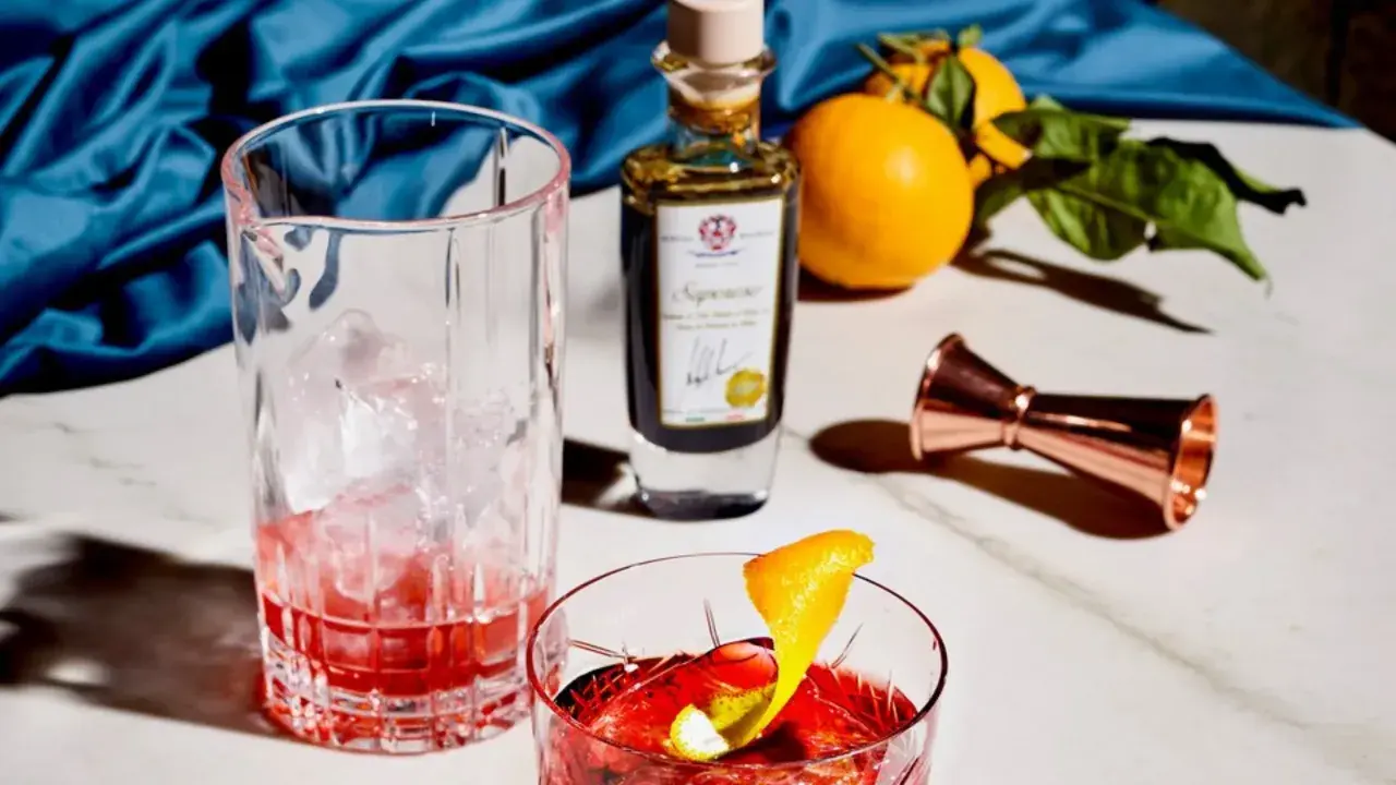 Ein Glas mit Eis und rotem Drink, daneben eine Flasche mit der Aufschrift "Saponoso". Orangen und ein Messbecher vervollständigen die Szene. Grappa, was ist das?