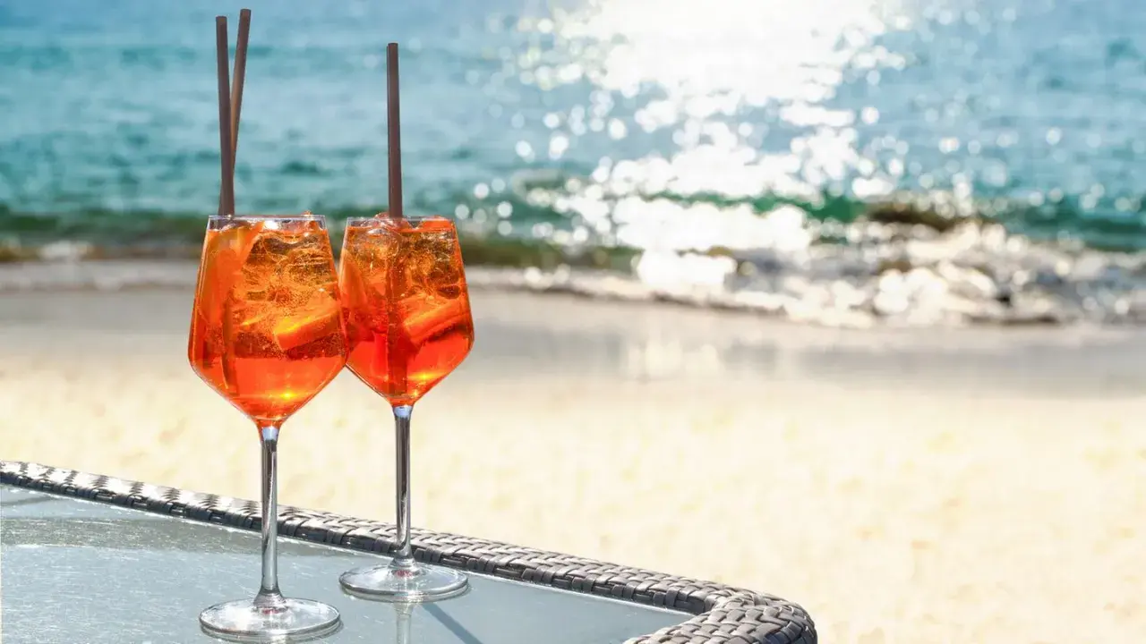 Zwei Aperol Spritz Gläser mit Eis und Orangenscheiben stehen am Strand. Der Aperol alkoholgehalt ist perfekt für einen warmen Tag.