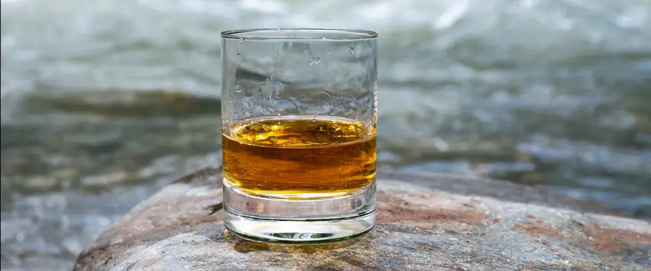 Ein Glas mit bernsteinfarbenem Whisky steht auf einem Stein. Im Hintergrund fließt ein Fluss.