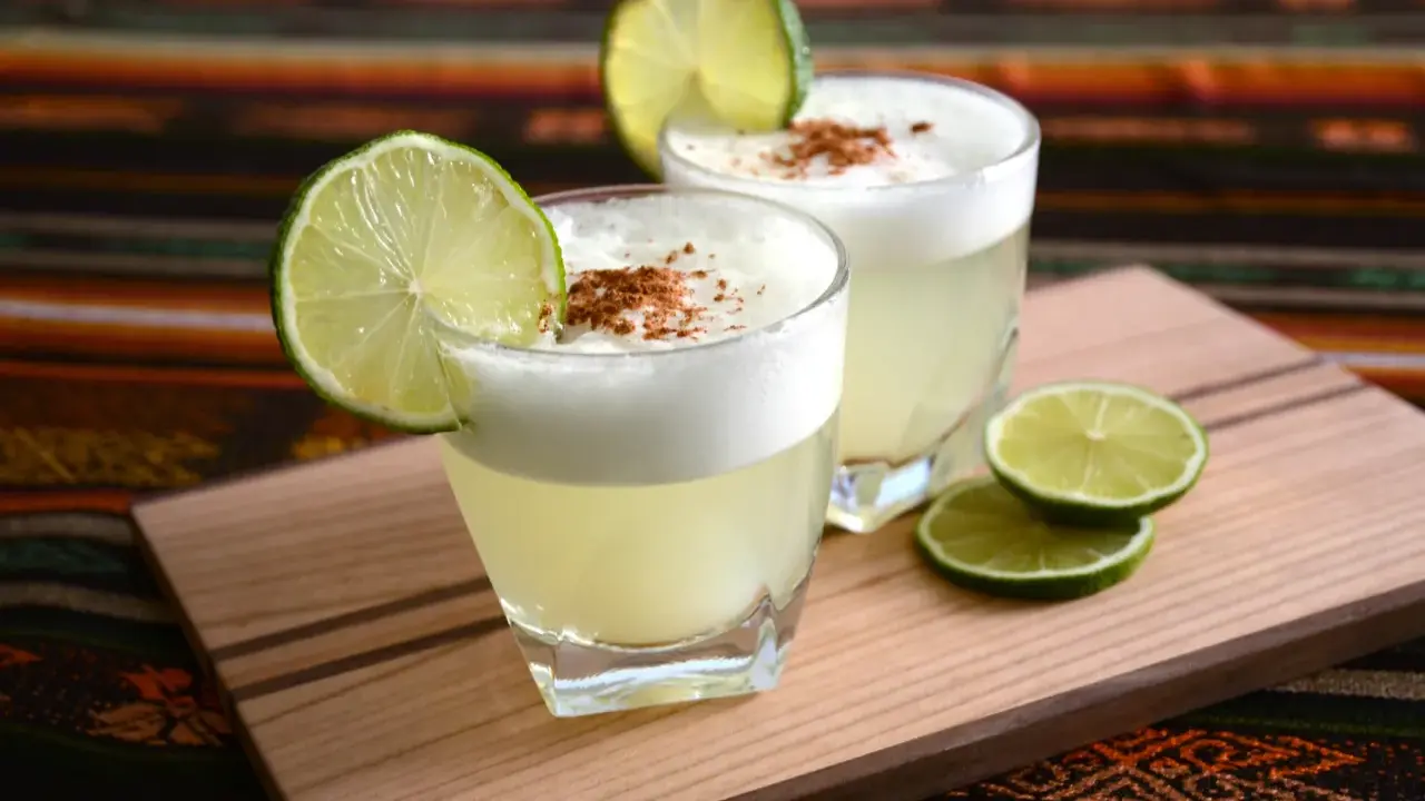 Zwei Pisco Sours, garniert mit Limettenspalten und Zimt, auf einem Holzbrett. Was ist Pisco? Ein peruanischer Traubenschnaps.