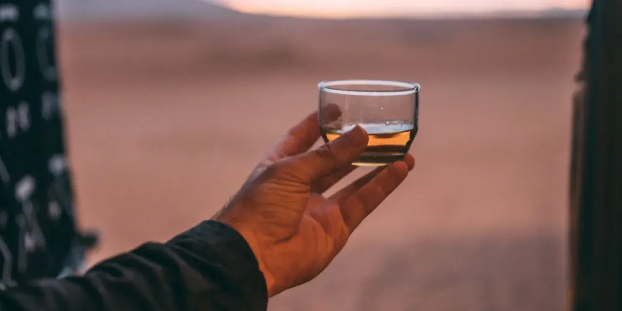 Hand hält ein Glas mit bernsteinfarbenem Rum verschnitt vor einer Wüstenlandschaft bei Sonnenuntergang.