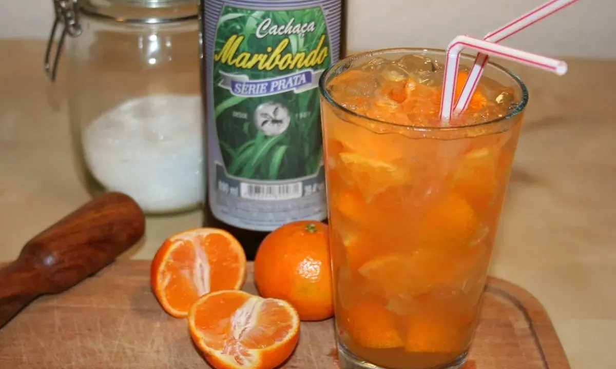 Ein Glas Caipirinha mit frischen Mandarinen, Eis und zwei Strohhalmen. Daneben eine Flasche Cachaça Maribondo, Zucker und ein Stößel.