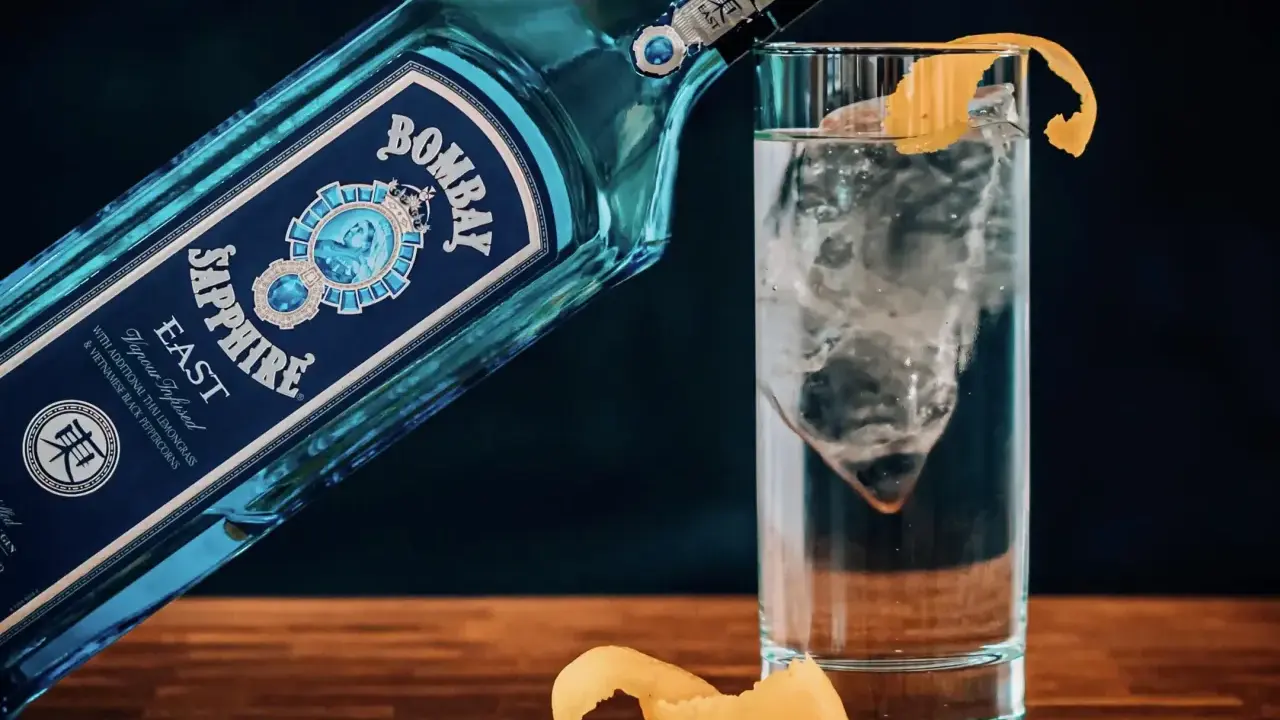 Eine Flasche Bombay Sapphire East Gin lehnt an einem Glas mit Eis und Zitronenschale. Was bedeutet Gin? Erfrischend und aromatisch.