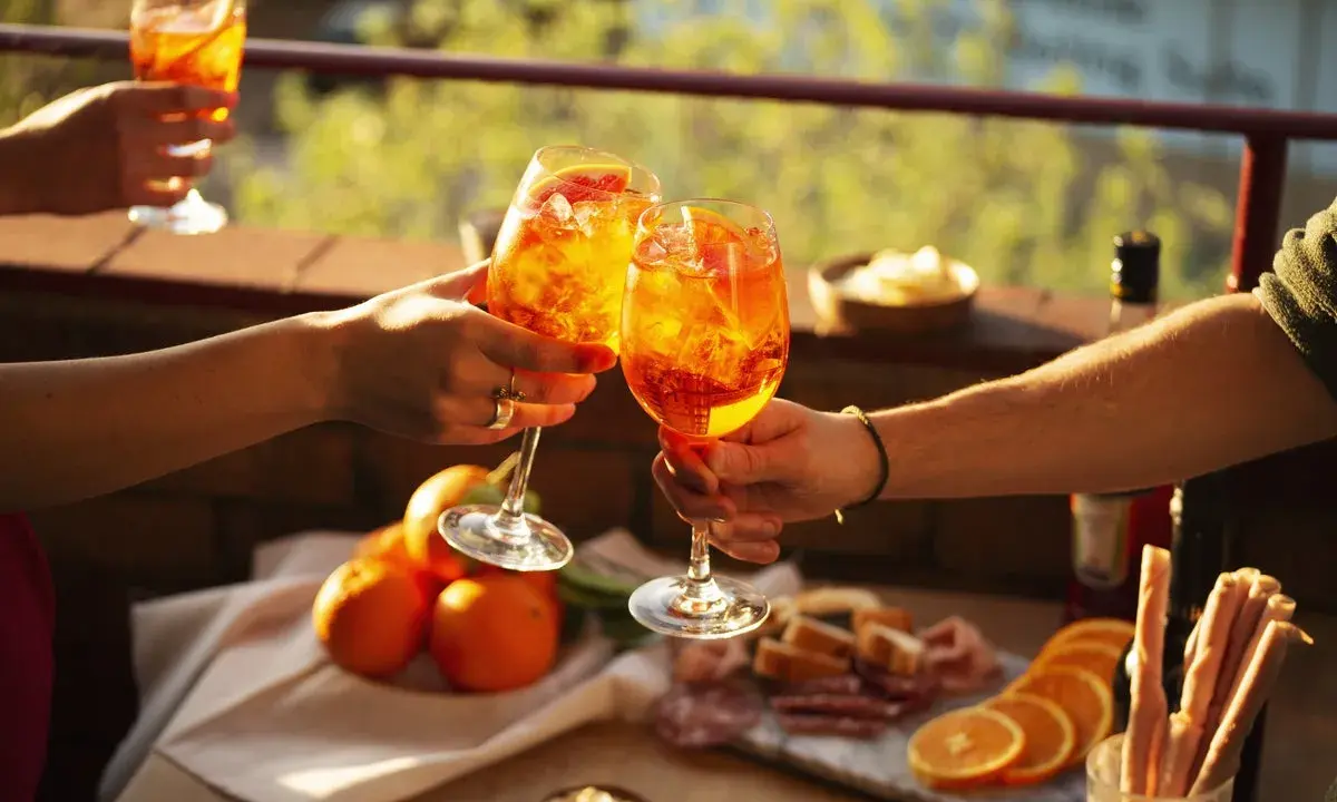 Zwei Hände stoßen mit Aperol-Cocktails an. Der Aperol, aus Rhabarber, Enzian, Chinarinde und Bitterorangen, leuchtet in der Sonne.