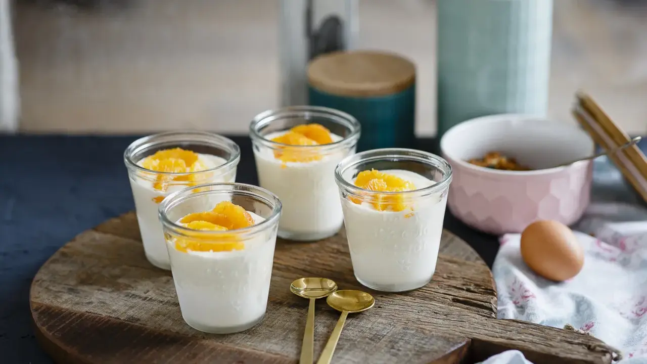 Vier Gläser mit cremigem Dessert, garniert mit Orangenspalten, ideal für Cointreau Rezepte. Zwei goldene Löffel liegen daneben.