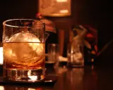 Ein Glas bernsteinfarbener Whisky mit einem großen Eisball. Im Hintergrund sind weitere Gläser und eine gedämpfte Beleuchtung zu sehen, die zum Genuss einlädt, wenn man Whisky mit Eis trinkt.