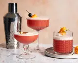 Drei erfrischende Campari Sours, garniert mit Orange und Kirsche, neben einem Cocktail-Shaker.