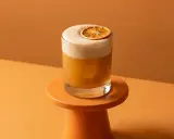 Ein cremiger Amaretto Sour mit einer getrockneten Orangenscheibe auf einem kleinen orangefarbenen Podest, vor einem warmen, orangenen Hintergrund.