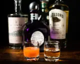 Zwei Gläser mit Tequila und einem orangen Getränk, bereit, um sangrita selber machen zu üben. Flaschen im Hintergrund.