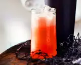 Erfrischender Aperol Sour mit Eiswürfeln in einem Glas, daneben ein dunkler Becher und Spinnweben-Deko.