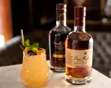 Ein erfrischender Cocktail mit Minze und Früchten neben zwei Flaschen Dos Maderas Rum. Der Alkoholgehalt des Rums ist auf dem Etikett angegeben.