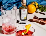 Ein Glas mit Eis und rotem Drink, daneben eine Flasche mit der Aufschrift "Saponoso". Orangen und ein Messbecher vervollständigen die Szene. Grappa, was ist das?