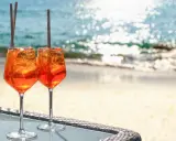 Zwei Aperol Spritz Gläser mit Eis und Orangenscheiben stehen am Strand. Der Aperol alkoholgehalt ist perfekt für einen warmen Tag.