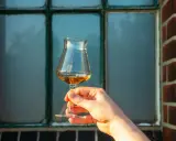 Hand hält ein Glas bernsteinfarbenen Whiskys vor einem Fenster. Der Alkoholgehalt variiert, aber oft liegt er bei 40 Prozent.