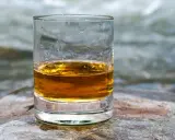 Ein Glas mit bernsteinfarbenem Whisky steht auf einem Stein. Im Hintergrund fließt ein Fluss.