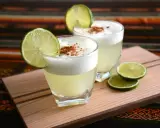 Zwei Pisco Sours, garniert mit Limettenspalten und Zimt, auf einem Holzbrett. Was ist Pisco? Ein peruanischer Traubenschnaps.