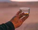 Hand hält ein Glas mit bernsteinfarbenem Rum verschnitt vor einer Wüstenlandschaft bei Sonnenuntergang.