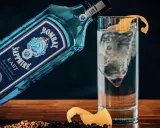 Eine Flasche Bombay Sapphire East Gin lehnt an einem Glas mit Eis und Zitronenschale. Was bedeutet Gin? Erfrischend und aromatisch.