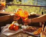 Zwei Hände stoßen mit Aperol-Cocktails an. Der Aperol, aus Rhabarber, Enzian, Chinarinde und Bitterorangen, leuchtet in der Sonne.