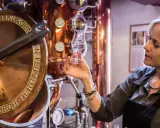 Frau verkostet Genever in einer Brennerei. Kupferne Anlagen und ein Glas mit dem klaren Schnaps sind zu sehen.