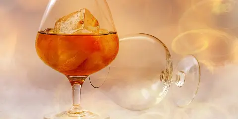 Ein Glas bernsteinfarbener Brandy mit Eiswürfeln, daneben ein leeres, umgekipptes Glas. Man fragt sich, wie schmeckt brandy.