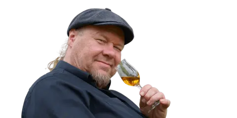 Ein Mann mit Schiebermütze und Bart hält ein Glas bernsteinfarbenen Rum. Er scheint zu wissen, wie man Rum genießt.