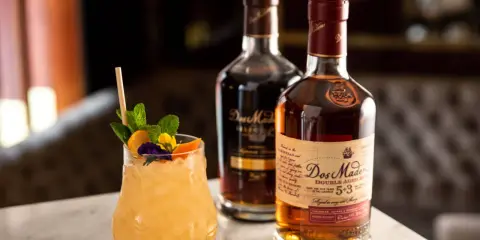 Ein erfrischender Cocktail mit Minze und Früchten neben zwei Flaschen Dos Maderas Rum. Der Alkoholgehalt des Rums ist auf dem Etikett angegeben.