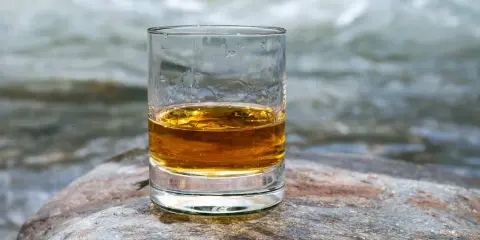 Ein Glas mit bernsteinfarbenem Whisky steht auf einem Stein. Im Hintergrund fließt ein Fluss.