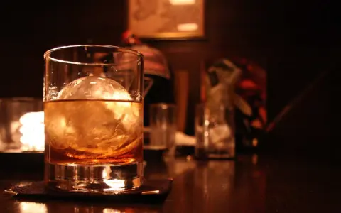 Ein Glas bernsteinfarbener Whisky mit einem großen Eisball. Im Hintergrund sind weitere Gläser und eine gedämpfte Beleuchtung zu sehen, die zum Genuss einlädt, wenn man Whisky mit Eis trinkt.