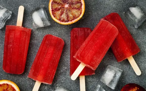 Erfrischende Eis am Stiel mit Campari Orange Geschmack, umgeben von Eiswürfeln und Blutorangen-Scheiben.