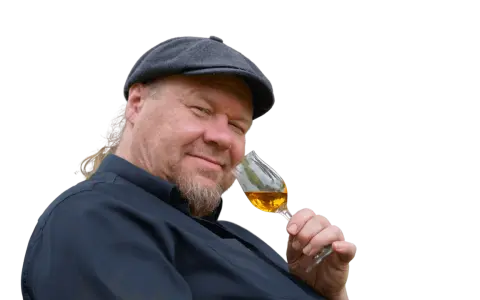 Ein Mann mit Schiebermütze und Bart hält ein Glas bernsteinfarbenen Rum. Er scheint zu wissen, wie man Rum genießt.
