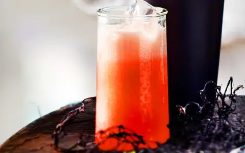 Erfrischender Aperol Sour mit Eiswürfeln in einem Glas, daneben ein dunkler Becher und Spinnweben-Deko.