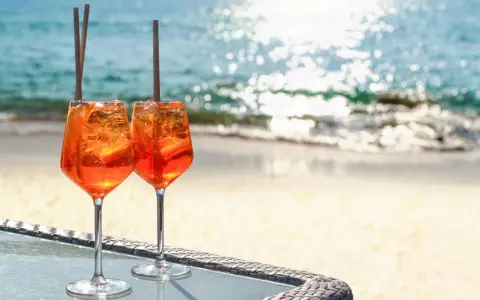 Zwei Aperol Spritz Gläser mit Eis und Orangenscheiben stehen am Strand. Der Aperol alkoholgehalt ist perfekt für einen warmen Tag.