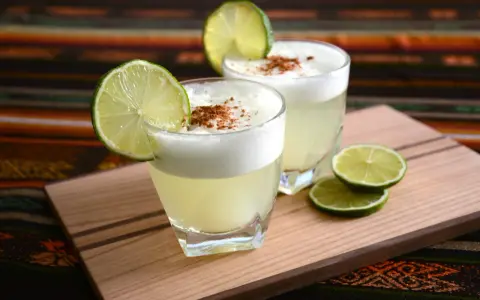 Zwei Pisco Sours, garniert mit Limettenspalten und Zimt, auf einem Holzbrett. Was ist Pisco? Ein peruanischer Traubenschnaps.