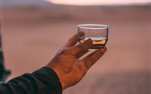 Hand hält ein Glas mit bernsteinfarbenem Rum verschnitt vor einer Wüstenlandschaft bei Sonnenuntergang.