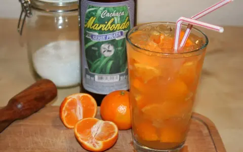Ein Glas Caipirinha mit frischen Mandarinen, Eis und zwei Strohhalmen. Daneben eine Flasche Cachaça Maribondo, Zucker und ein Stößel.