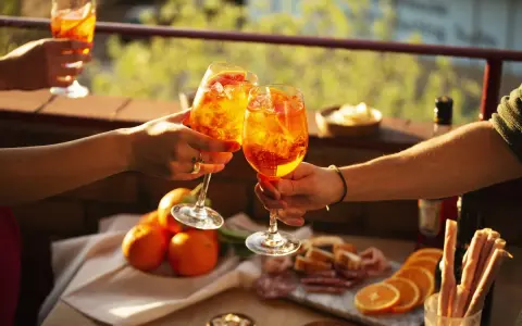Zwei Hände stoßen mit Aperol-Cocktails an. Der Aperol, aus Rhabarber, Enzian, Chinarinde und Bitterorangen, leuchtet in der Sonne.
