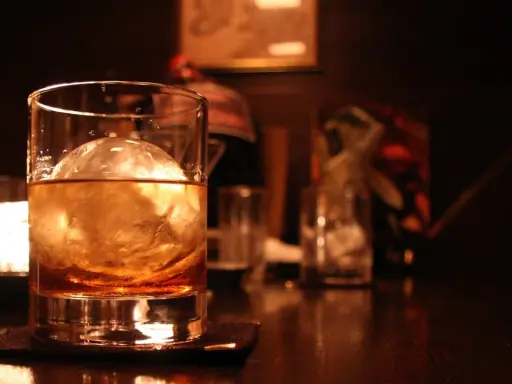 Ein Glas bernsteinfarbener Whisky mit einem großen Eisball. Im Hintergrund sind weitere Gläser und eine gedämpfte Beleuchtung zu sehen, die zum Genuss einlädt, wenn man Whisky mit Eis trinkt.