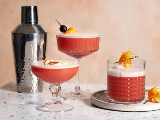 Drei erfrischende Campari Sours, garniert mit Orange und Kirsche, neben einem Cocktail-Shaker.