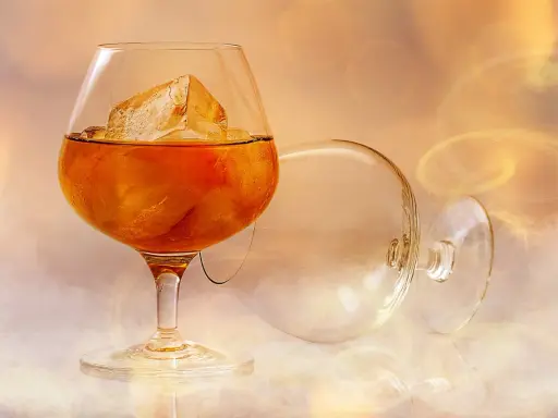 Ein Glas bernsteinfarbener Brandy mit Eiswürfeln, daneben ein leeres, umgekipptes Glas. Man fragt sich, wie schmeckt brandy.