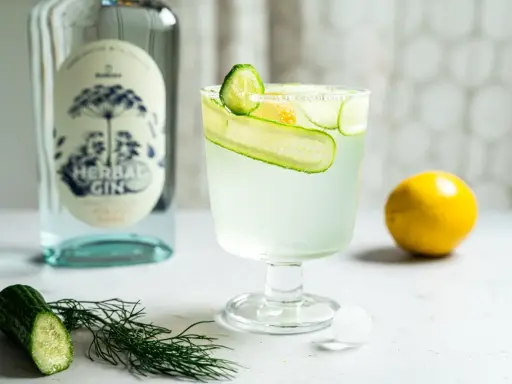 Erfrischender Gin Tonic mit Gurke und Zitrone, daneben eine Flasche Gin und frische Zutaten.