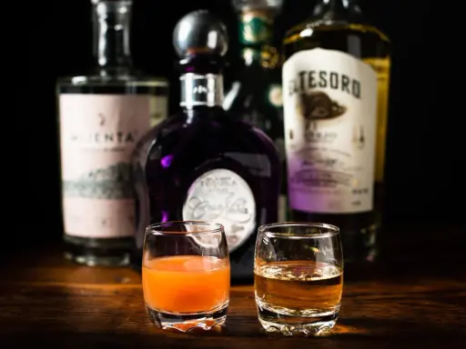 Zwei Gläser mit Tequila und einem orangen Getränk, bereit, um sangrita selber machen zu üben. Flaschen im Hintergrund.