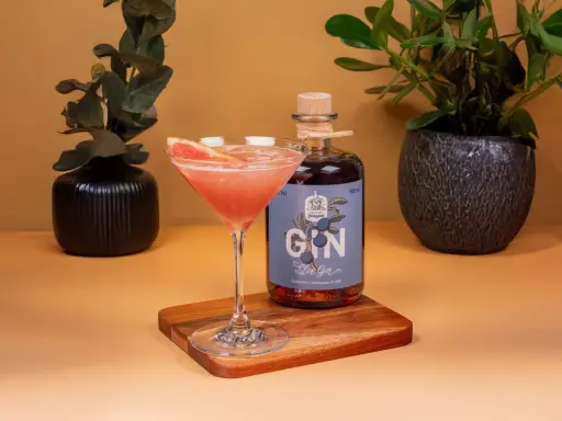 Ein Glas mit pinkfarbenem Gin-Cocktail, garniert mit Grapefruit, neben einer Flasche Sloe Gin. Dekorative Pflanzen im Hintergrund.