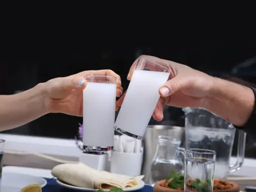 Zwei Hände stoßen mit Gläsern voller weißem Raki an. So trinkt man Raki: langsam, mit Freunden und gutem Essen.